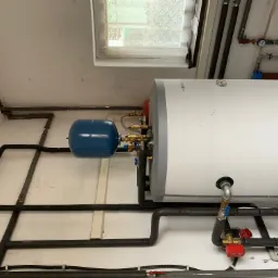 Instalacja pompy ciepła w kotłowni: biały zasobnik ciepłej wody, niebieskie naczynie przeponowe, rury w izolacji termicznej, okno w tle.