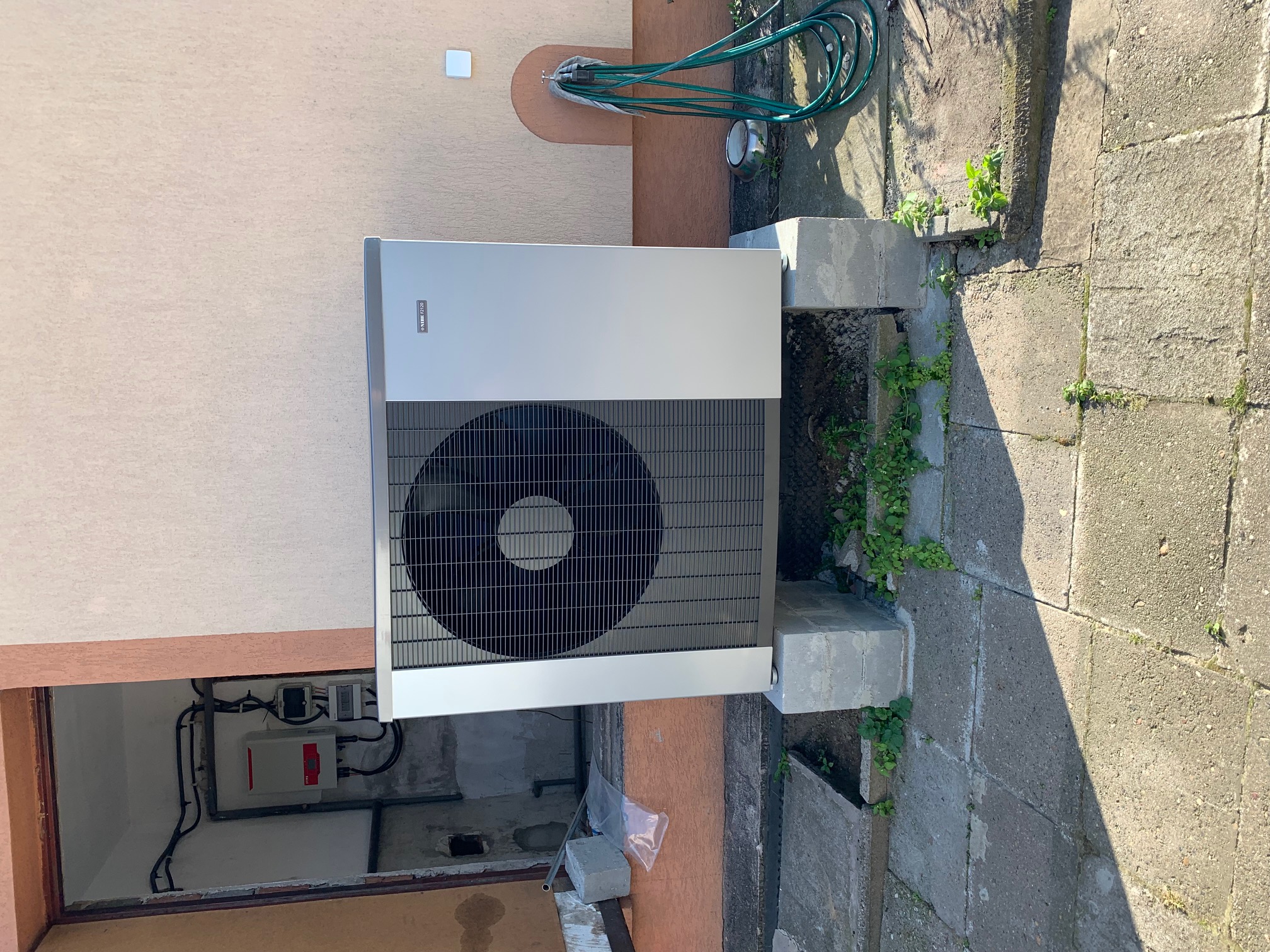 Zewnętrzna jednostka pompy ciepła marki NIBE ustawiona na betonowych bloczkach, widoczne podłączenia elektryczne i przewody, zielony wąż ogrodowy zwinięty na ścianie budynku