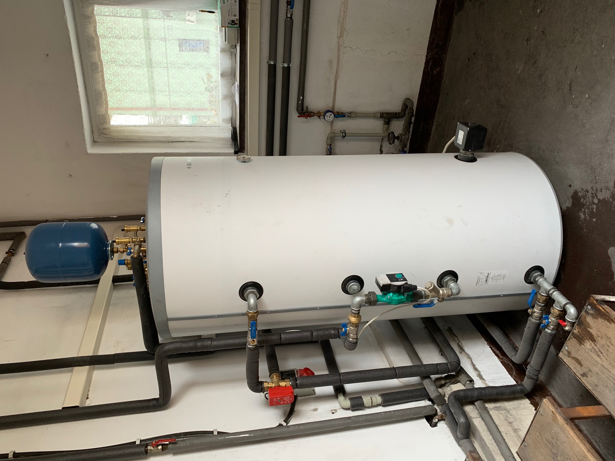 Biały akumulator ciepła z podłączonymi rurami w kotłowni w Przystajni, widoczny niebieski zbiornik wyrównawczy i okno z firanką w tle.