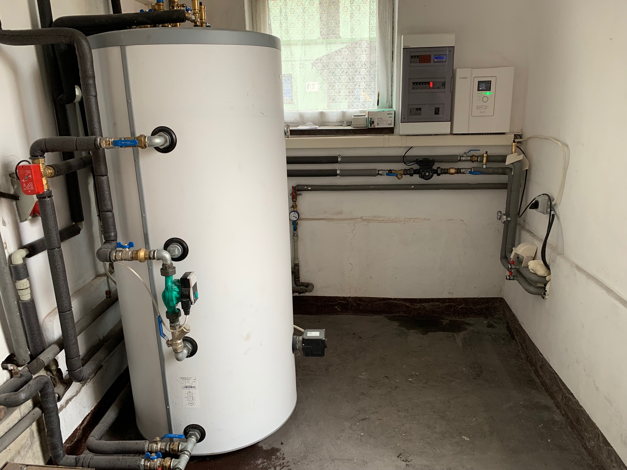 Biały zbiornik buforowy z licznymi rurami i zaworami w piwnicy, obok ściany z panelami sterowania i okienkiem z firanką.
