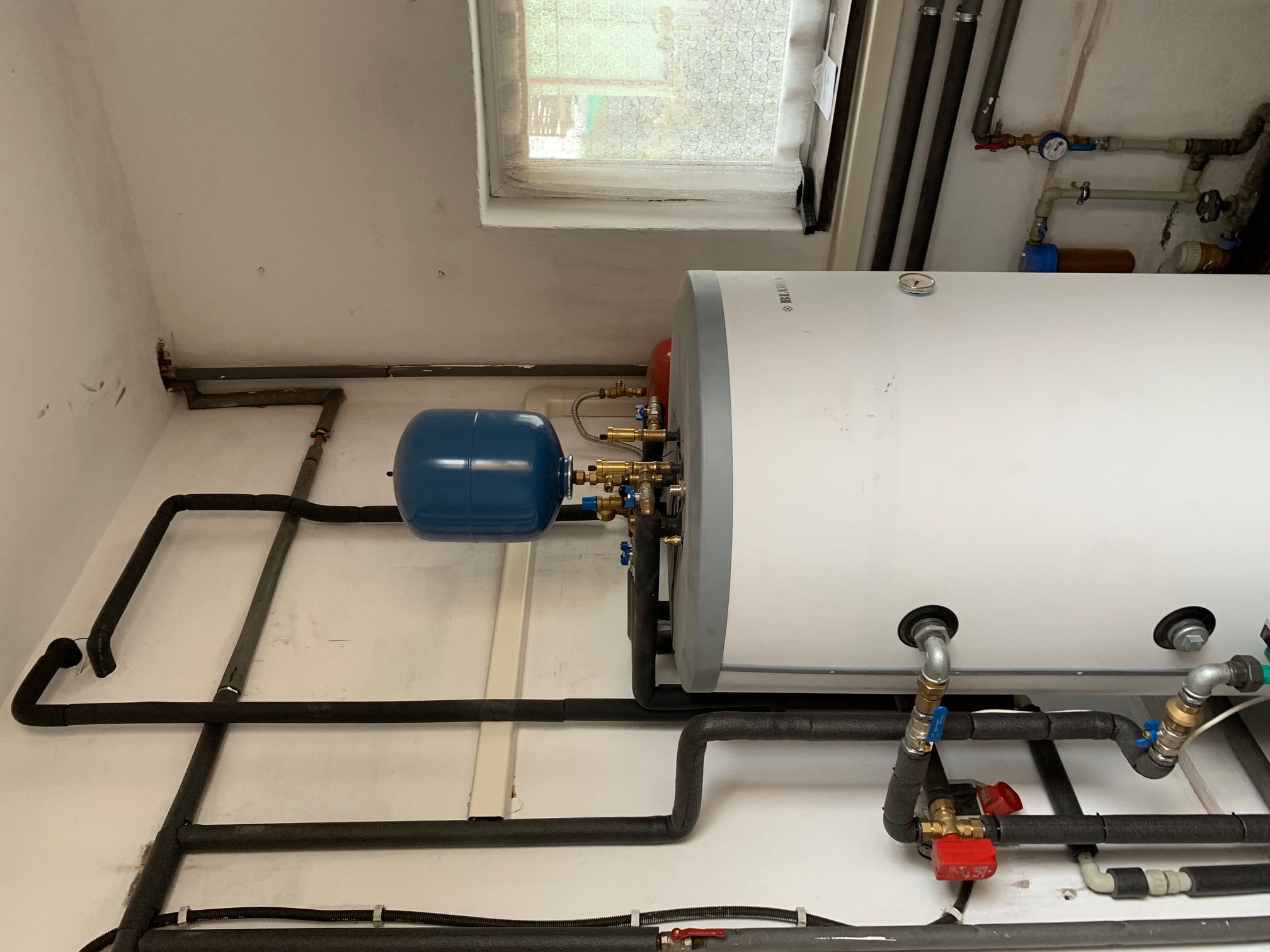 Instalacja pompy ciepła w kotłowni: biały zasobnik ciepłej wody, niebieskie naczynie przeponowe, rury w izolacji termicznej, okno w tle.