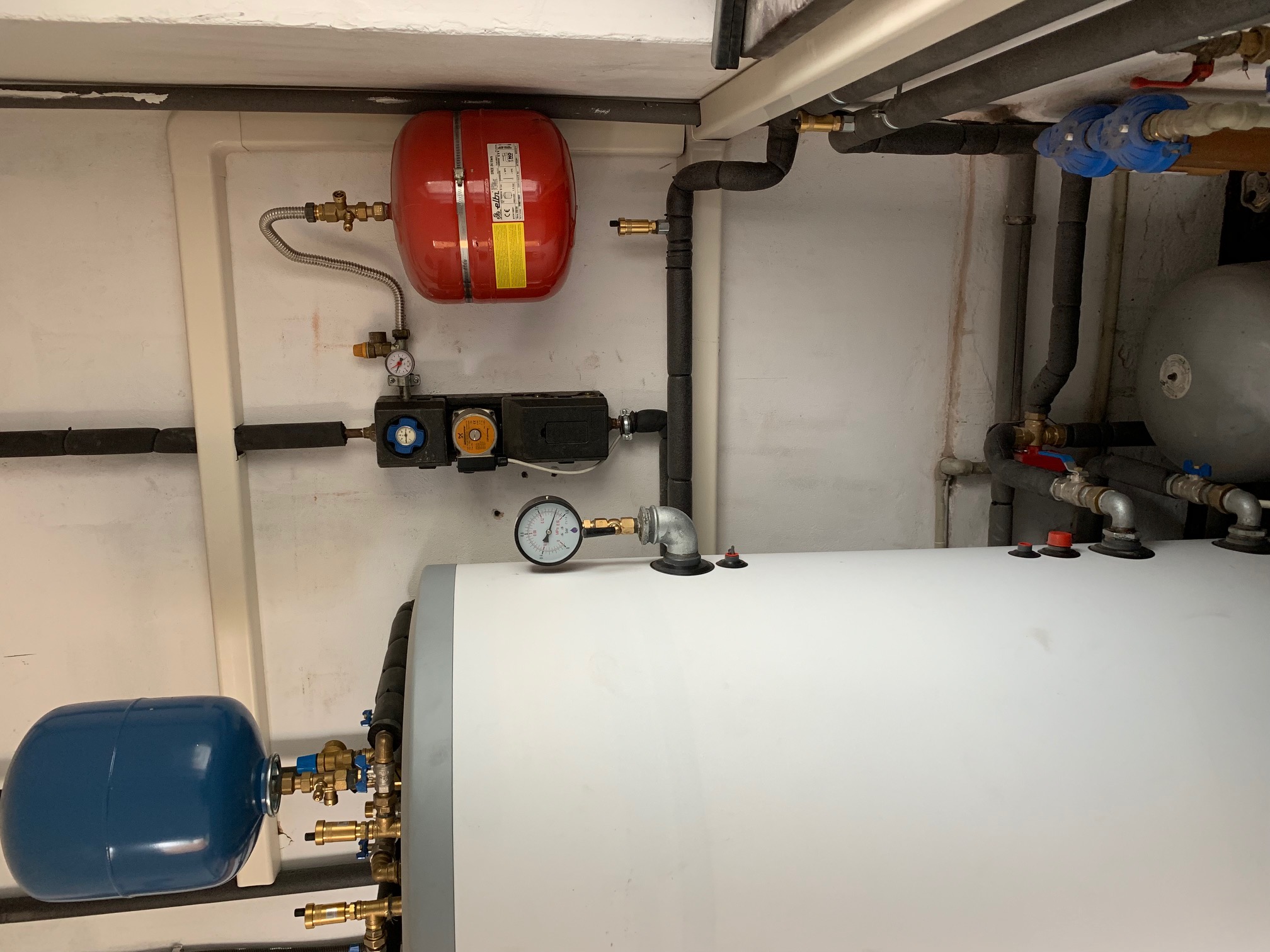 Instalacja grzewcza w kotłowni: biały zbiornik buforowy z manometrem, czerwony zbiornik wyrównawczy i niebieski zbiornik ciśnieniowy, połączone rurami z izolacją.