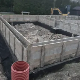 Deskowanie pod wylewkę fundamentów z widocznym zbrojeniem, rurą drenarską i koparką w tle.