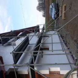 Ocieplanie elewacji budynku jednorodzinnego z użyciem rusztowania, widoczne płyty styropianowe i cegły, w tle samochód osobowy.