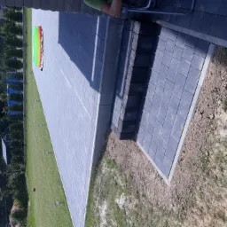 Nowoczesny taras z szarej kostki brukowej, z widocznymi schodami i fragmentem trawnika z dziecięcym basenem w tle.