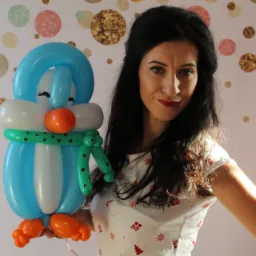 Uśmiechnięta brunetka prezentuje figurkę pingwina wykonaną z modelowanych balonów na tle ściany w kropki.