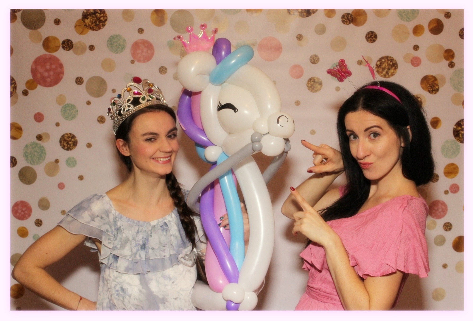 Dwie kobiety w kostiumach pozują z balonową figurą jednorożca na tle ściany w kropki. Jedna kobieta ma koronę, druga opaskę z motylkiem.