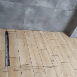 Ułożone płytki podłogowe imitujące drewno, z widocznym odpływem liniowym i szarą płytką ścienną w tle.