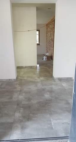 Wnętrze w trakcie prac wykończeniowych: szara, marmurkowa podłoga, surowe ściany w odcieniach bieli, fragment odsłoniętej cegły w głębi pomieszczenia, widoczne materiały budowlane.