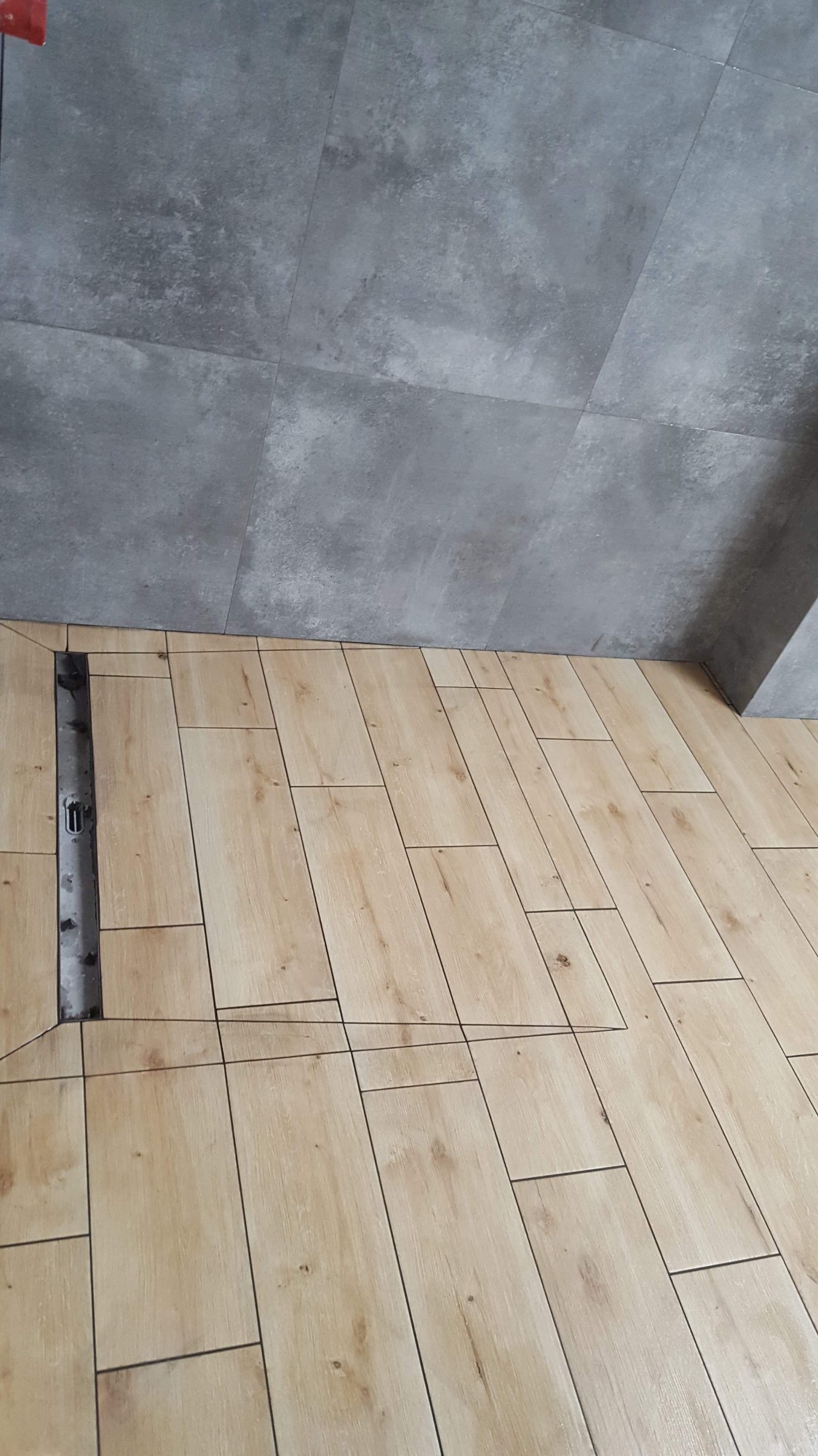 Ułożone płytki podłogowe imitujące drewno, z widocznym odpływem liniowym i szarą płytką ścienną w tle.