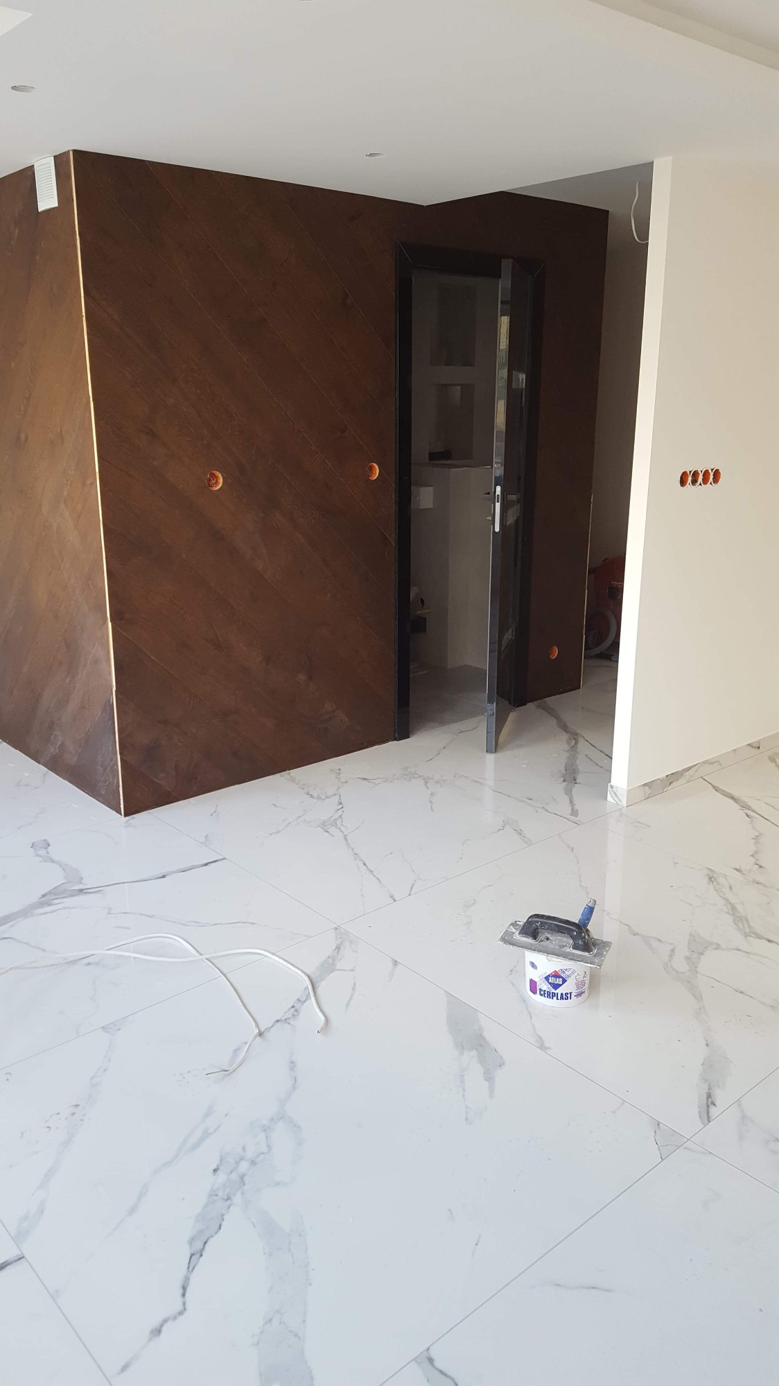 Wykończone wnętrze: ściana z drewnianą okładziną ułożoną w jodełkę, biała marmurowa podłoga, otwarte drzwi do łazienki, puszka z masą szpachlową i szpachelka na podłodze.