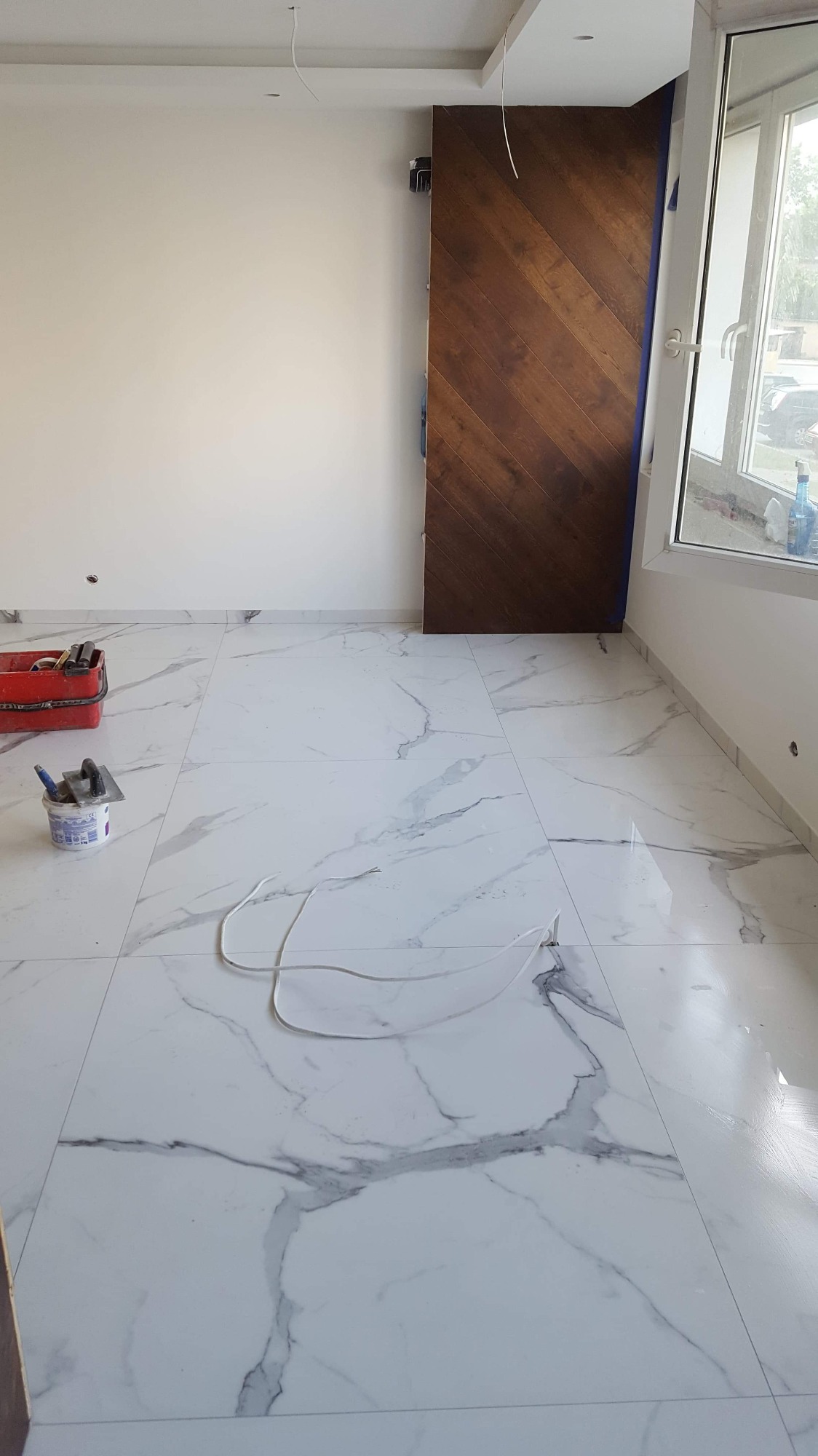 Pokój w trakcie wykańczania: białe marmurowe płytki podłogowe, drewniane drzwi przesuwne z wzorem jodełki, narzędzia i materiały budowlane w rogu, kabel na podłodze.