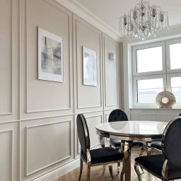 Roll and Wall Sebastian Ciszewski - Elegancka jadalnia w odcieniach beżu z panelami ściennymi, okrągłym stołem i kryształowym żyrandolem. Parkiet w jodełkę i abstrakcyjne obrazy dodają wnętrzu charakteru.