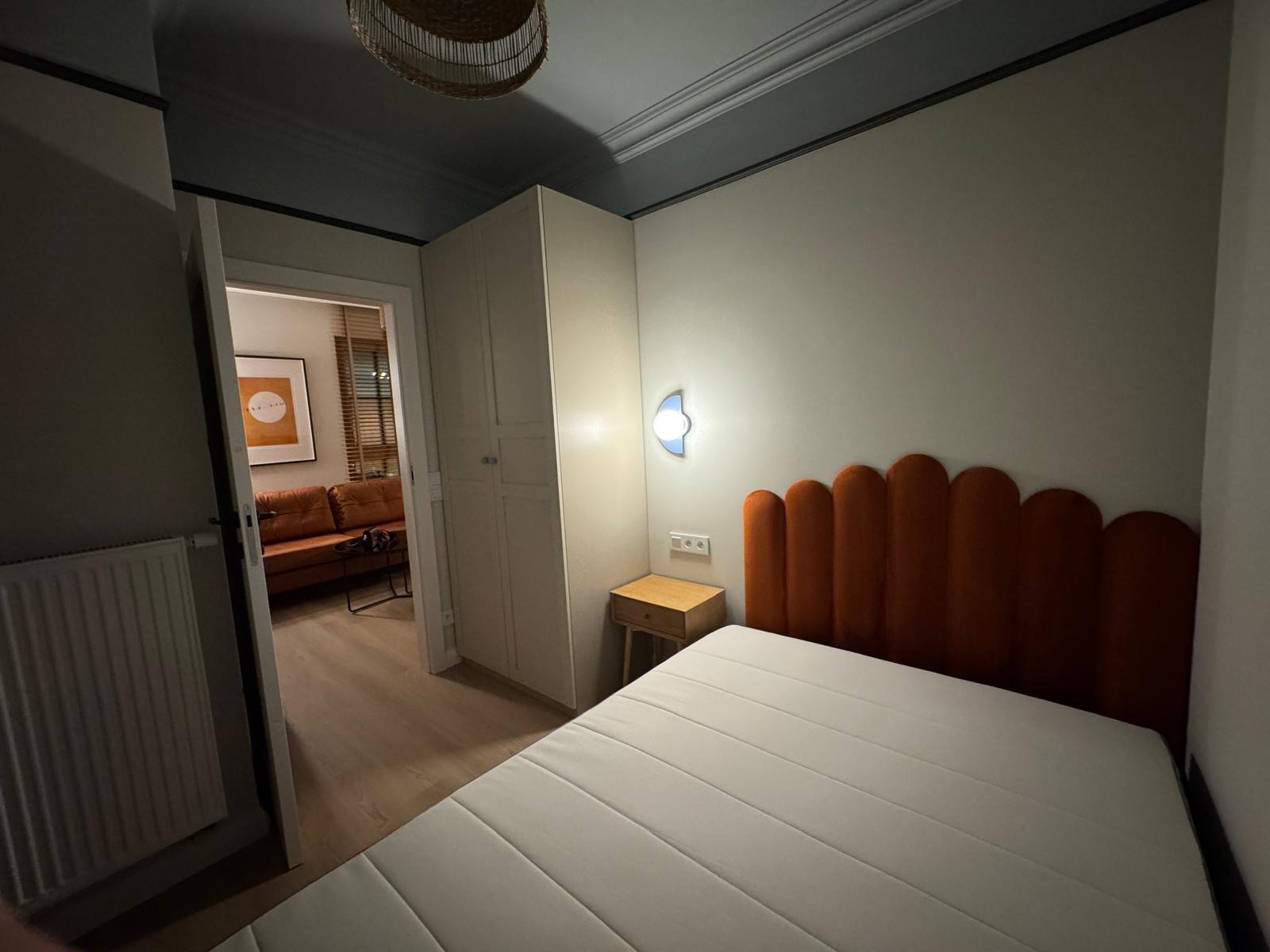 Nowoczesna sypialnia z łóżkiem z pomarańczowym wezgłowiem, szafką nocną i widokiem na salon przez otwarte drzwi. Minimalistyczny design, stonowana kolorystyka.