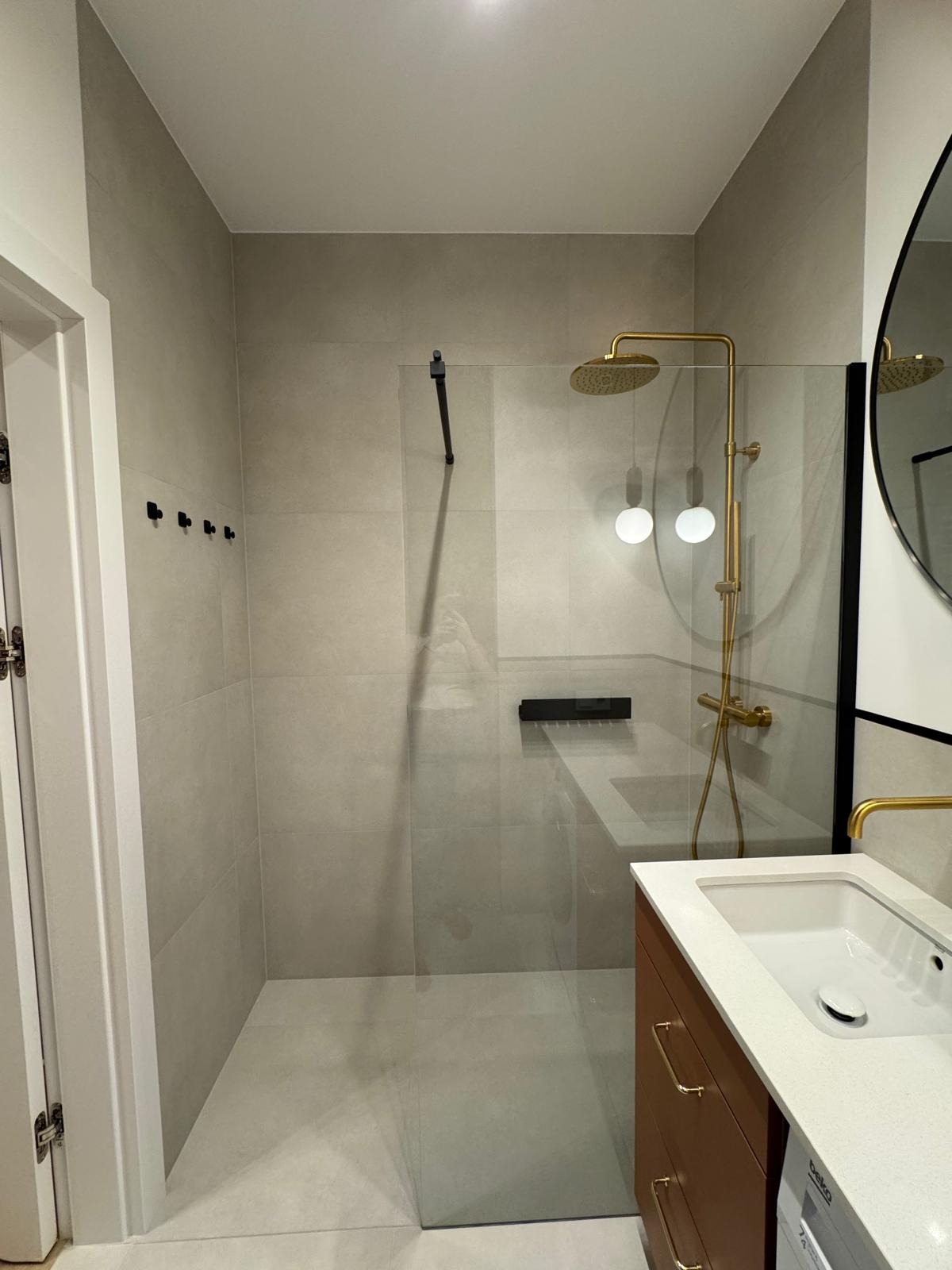 Nowoczesna łazienka z kabiną prysznicową typu walk-in, złotą armaturą i umywalką z szafką w kolorze brązowym. Minimalistyczny design, jasne płytki i czarne akcenty.