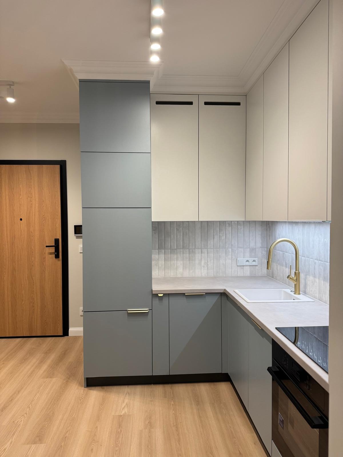 Nowoczesna kuchnia w odcieniach szarości i bieli z złotą armaturą. Minimalistyczny design, drewniana podłoga i drzwi. Oświetlenie sufitowe punktowe.