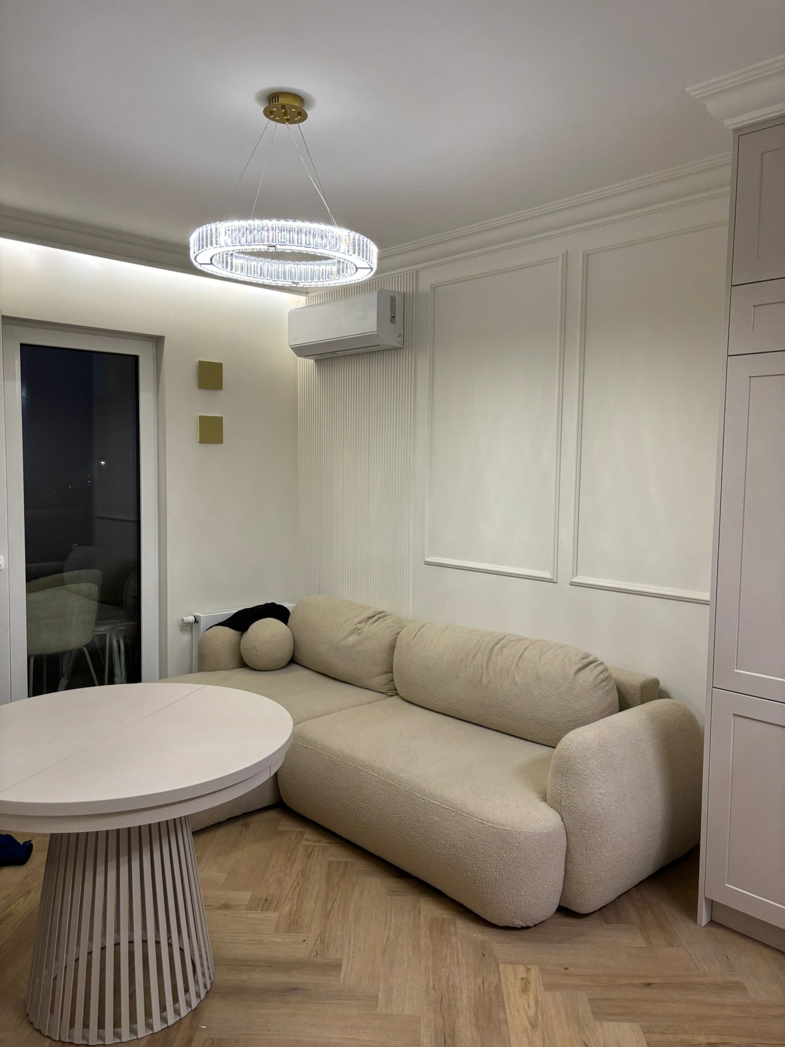 Jasne wnętrze: beżowa sofa z okrągłym stolikiem na parkiecie, ozdobne listwy na ścianie, nowoczesny żyrandol i klimatyzacja. Minimalistyczny design.