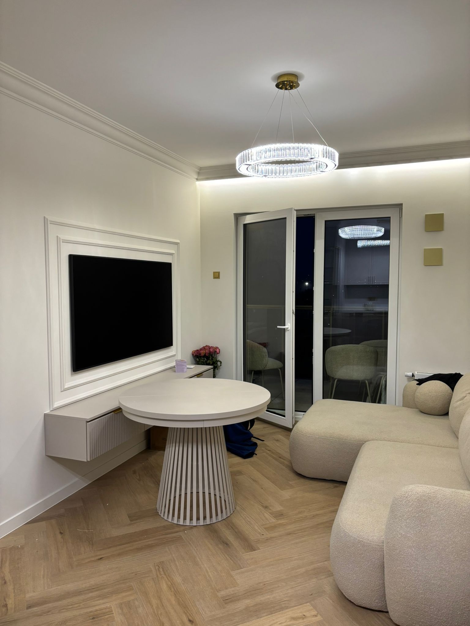 Eleganckie wykończenie wnętrza w Warszawie: salon z telewizorem w ozdobnej ramie, okrągły stół i jasna sofa. Parkiet w jodełkę i nowoczesna lampa.