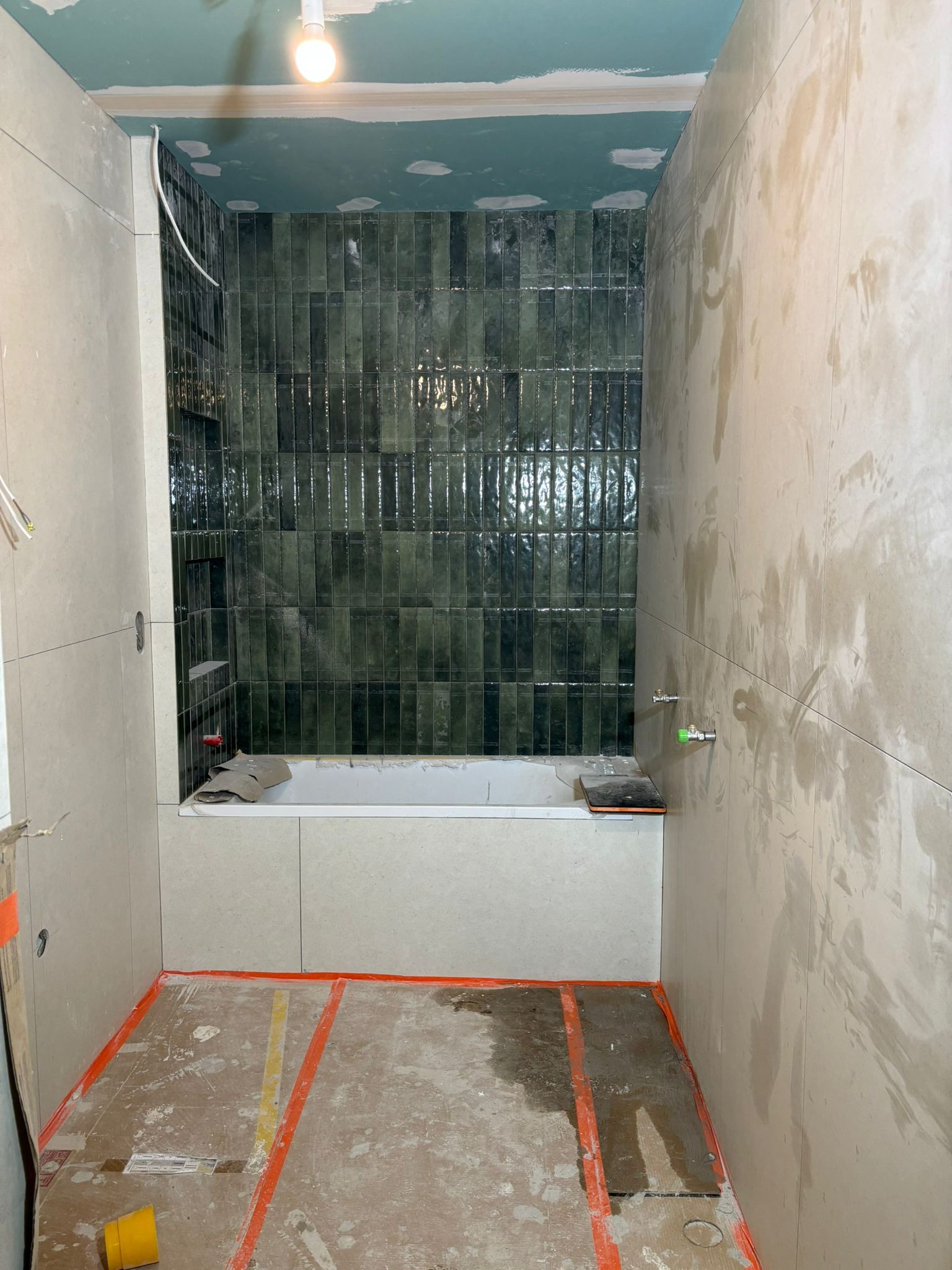 Remont łazienki w trakcie: zielona, pionowa glazura nad wanną, jasne płytki na ścianach, zabezpieczona podłoga. Widoczne elementy instalacji i niedokończone wykończenie.