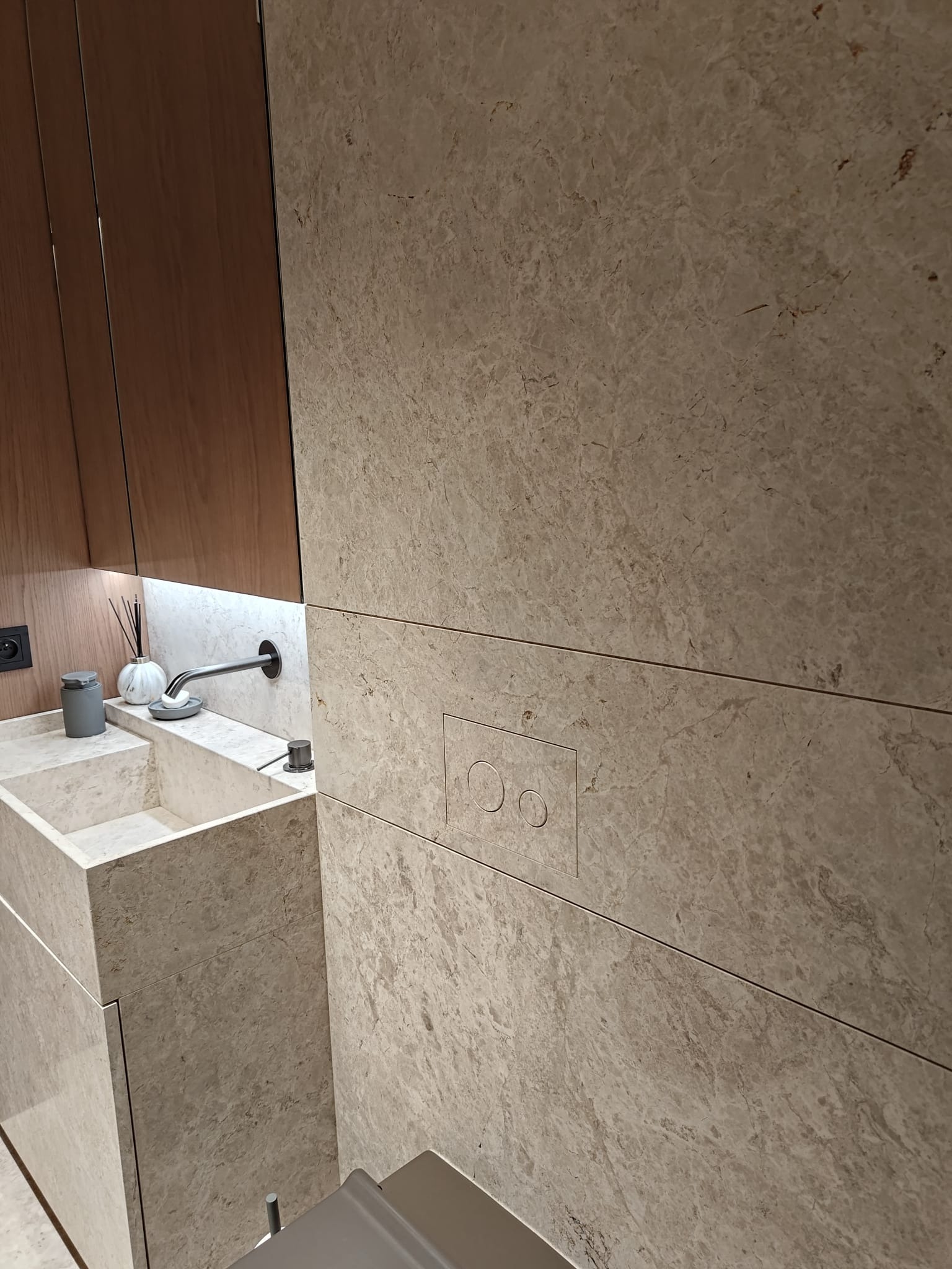 Beżowa łazienka z umywalką z kamienia i ukrytym panelem WC. Minimalistyczny design, kamienne wykończenie ścian i szafki pod umywalką. Nowoczesne oświetlenie.