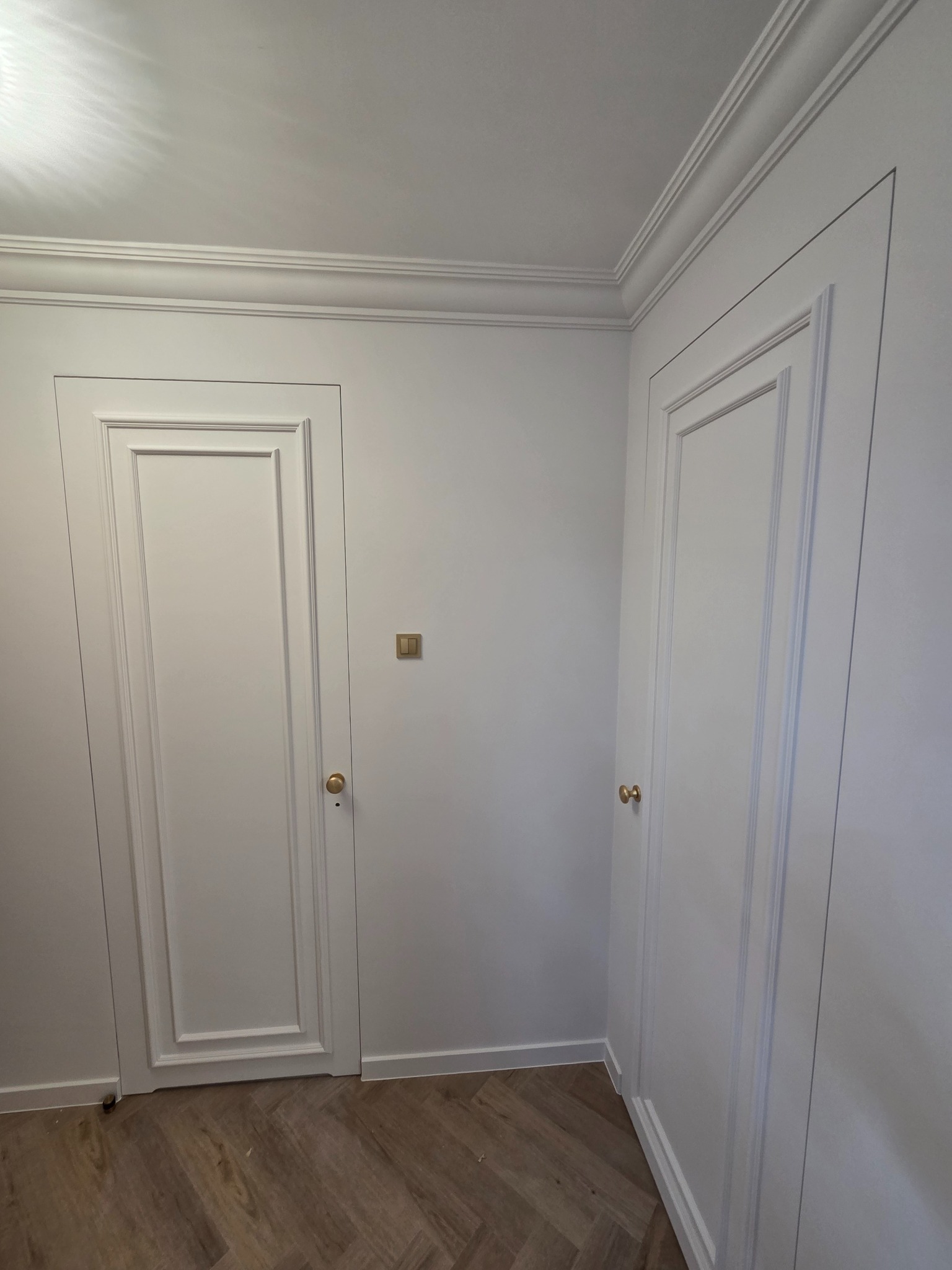 Białe, klasyczne drzwi wewnętrzne z ozdobnymi listwami i złotymi gałkami, wkomponowane w białe ściany z dekoracyjnym gzymsem sufitowym. Parkiet ułożony w jodełkę.