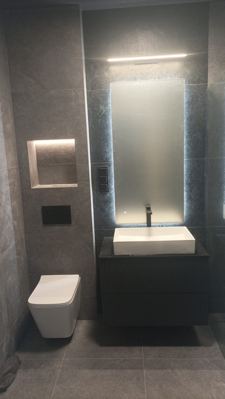 Nowoczesna łazienka w szarościach: podświetlane lustro, wisząca toaleta i umywalka nablatowa na szafce z ryflowanym frontem. Minimalistyczny design z akcentami światła.