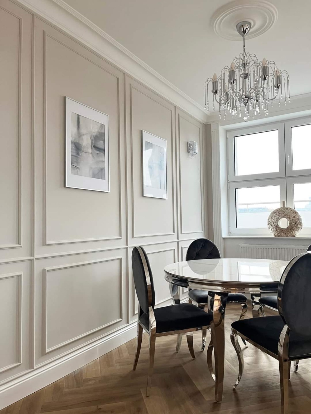 Elegancka jadalnia w odcieniach beżu z panelami ściennymi, okrągłym stołem i kryształowym żyrandolem. Parkiet w jodełkę i abstrakcyjne obrazy dodają wnętrzu charakteru.