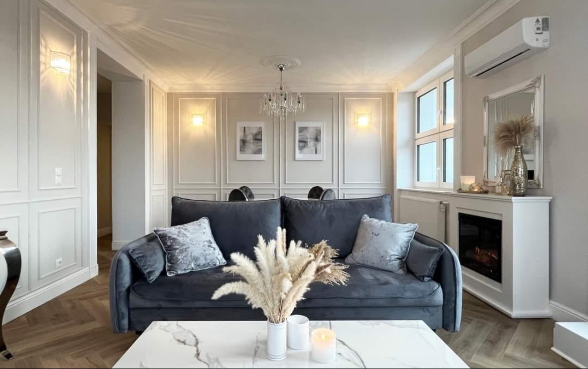 Elegancki salon w odcieniach szarości i bieli. Dekoracyjne panele ścienne, kryształowy żyrandol i marmurowy stolik kawowy tworzą luksusową atmosferę.