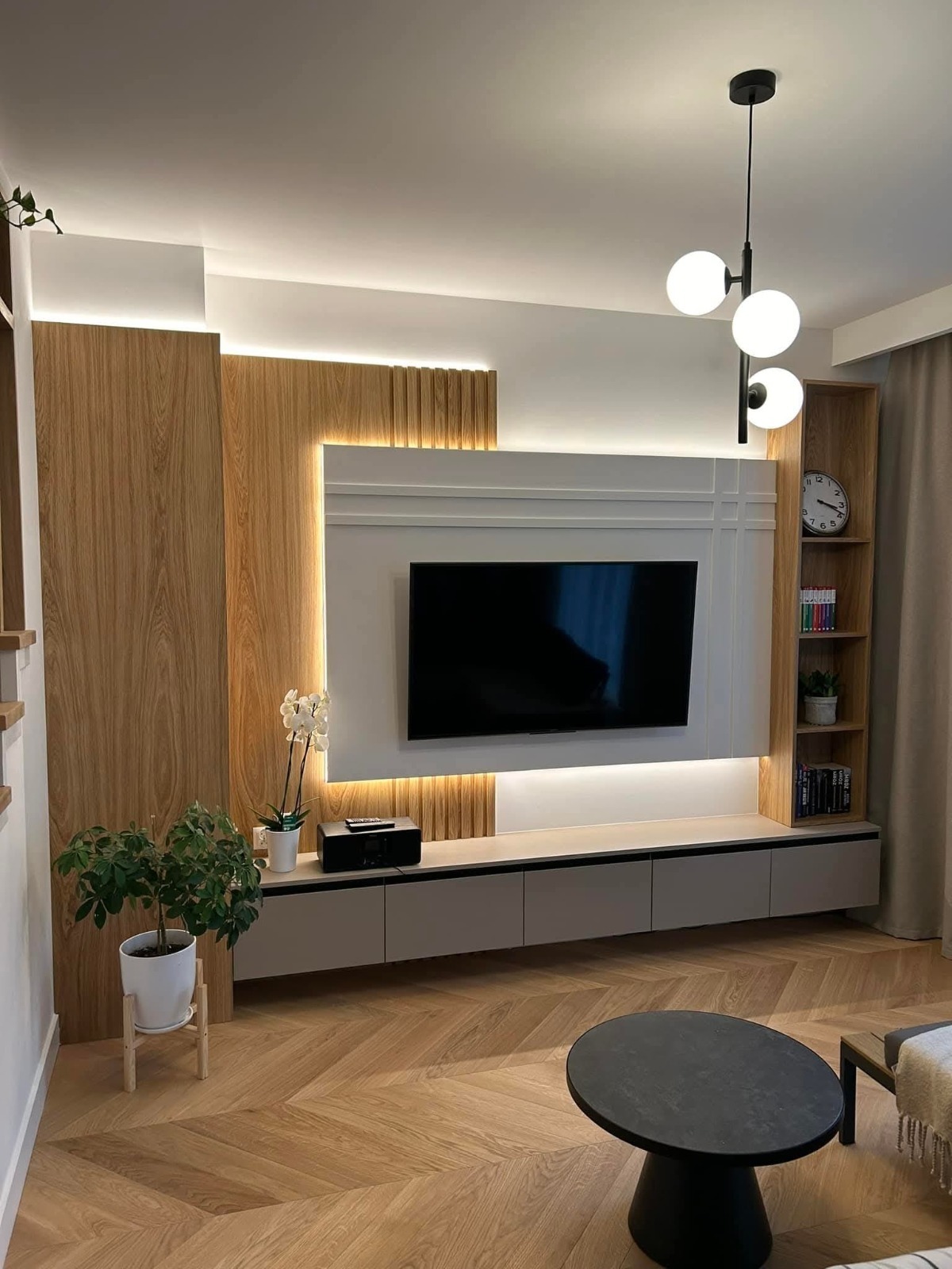 Nowoczesny salon z drewnianą ścianą TV, podświetleniem LED i parkietem w jodełkę. Minimalistyczny stolik kawowy i designerska lampa wisząca.