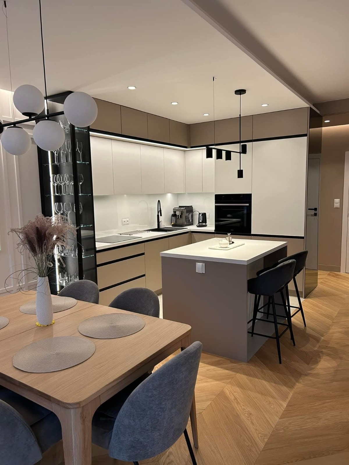 Nowoczesna kuchnia z wyspą i barkiem, oświetlona lampami wiszącymi. Parkiet ułożony w jodełkę. Szklana witryna z podświetleniem eksponuje kolekcję kieliszków.