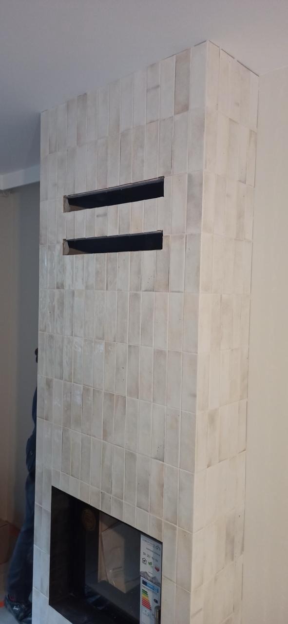 Obudowa kominka wykończona beżowymi, pionowo ułożonymi płytkami. Widoczne dwa otwory wentylacyjne i wkład kominkowy. Minimalistyczny design.