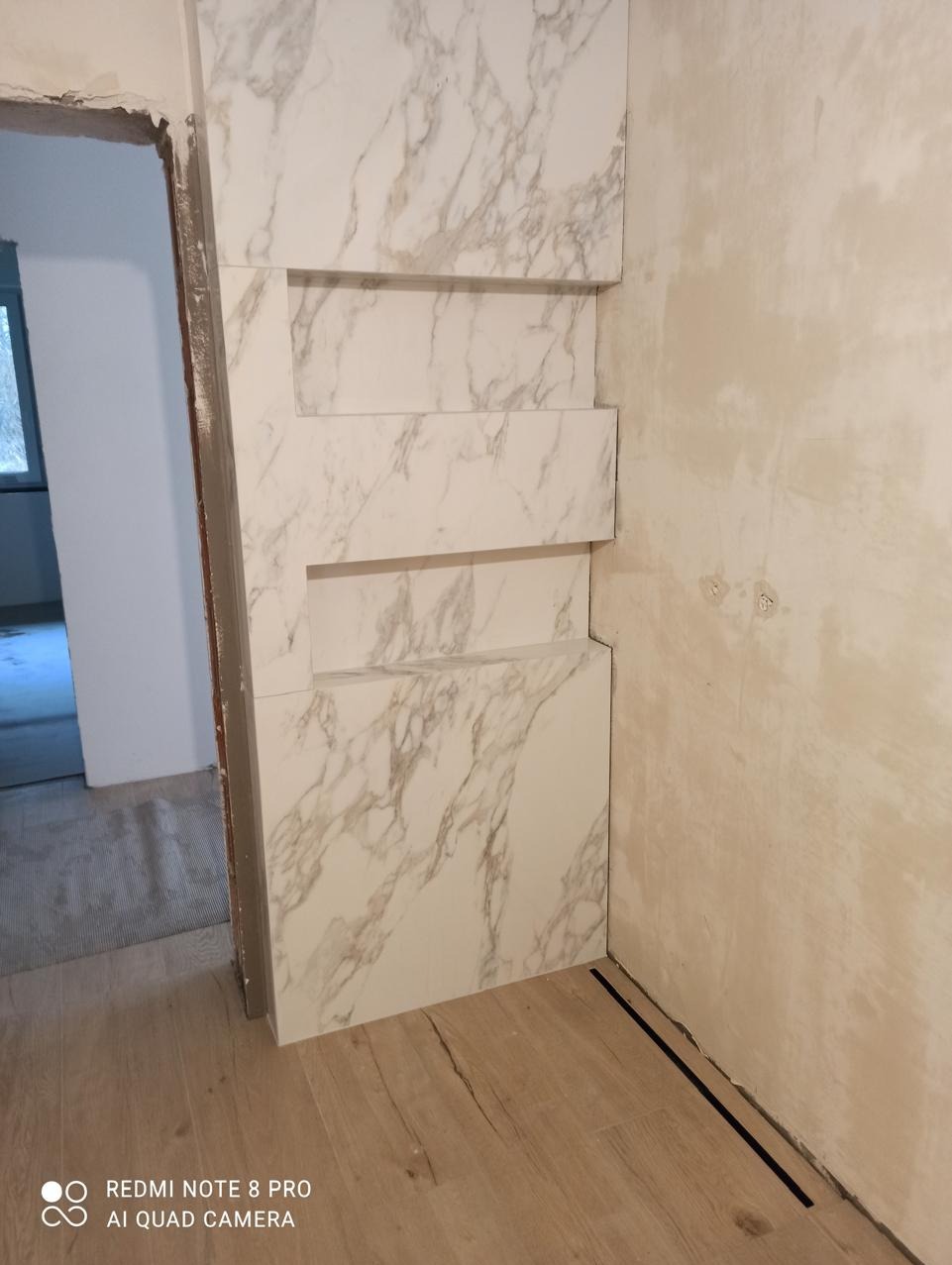 Ściana wyłożona płytkami imitującymi marmur z wnękami, widoczne niedokończone prace na sąsiedniej ścianie i drewnopodobna podłoga. Ujęcie z dołu.