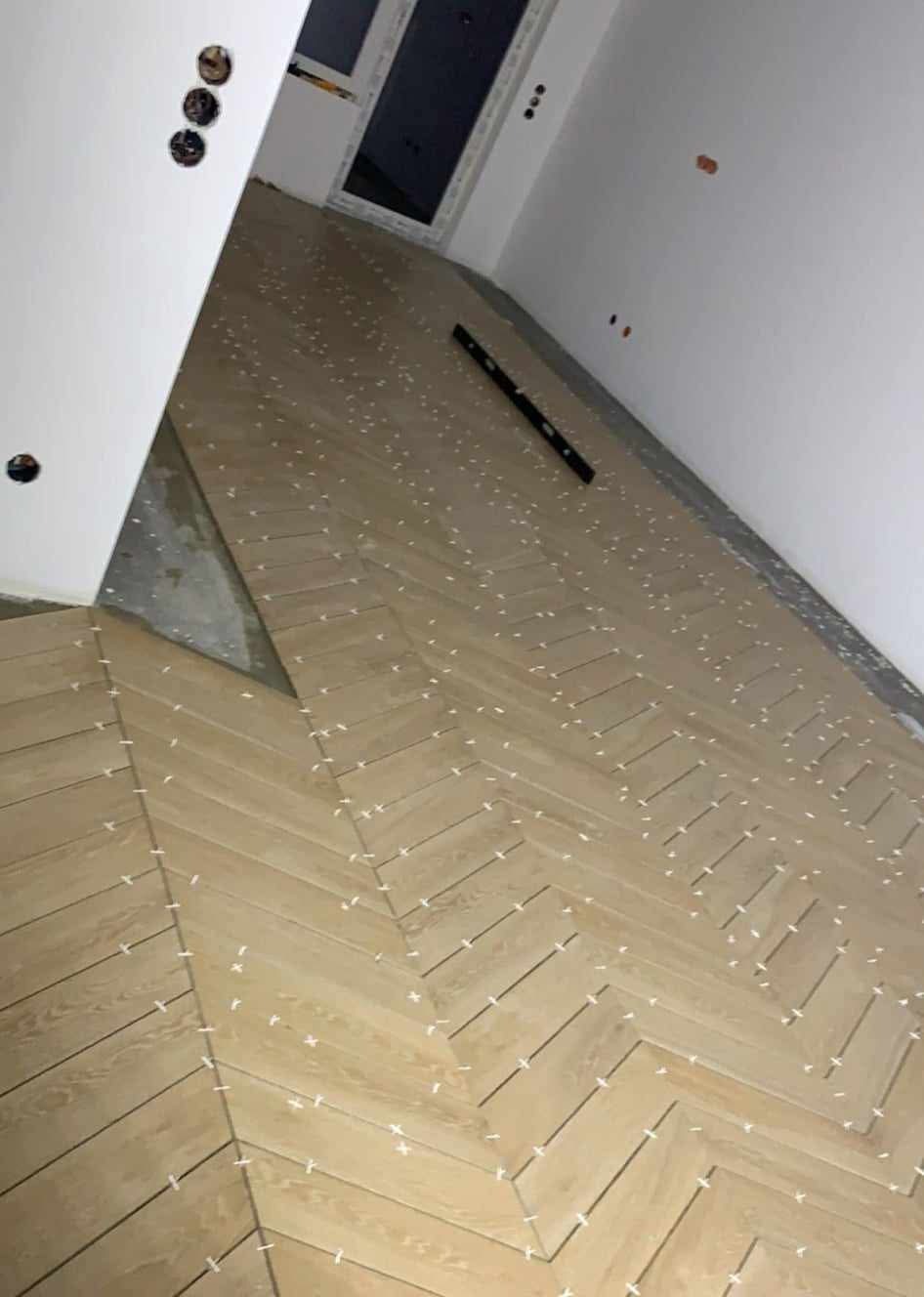 Układanie parkietu w jodełkę z użyciem białych krzyżyków dystansowych. Poziomica leży na podłodze. Widoczne gniazdka elektryczne na ścianie.