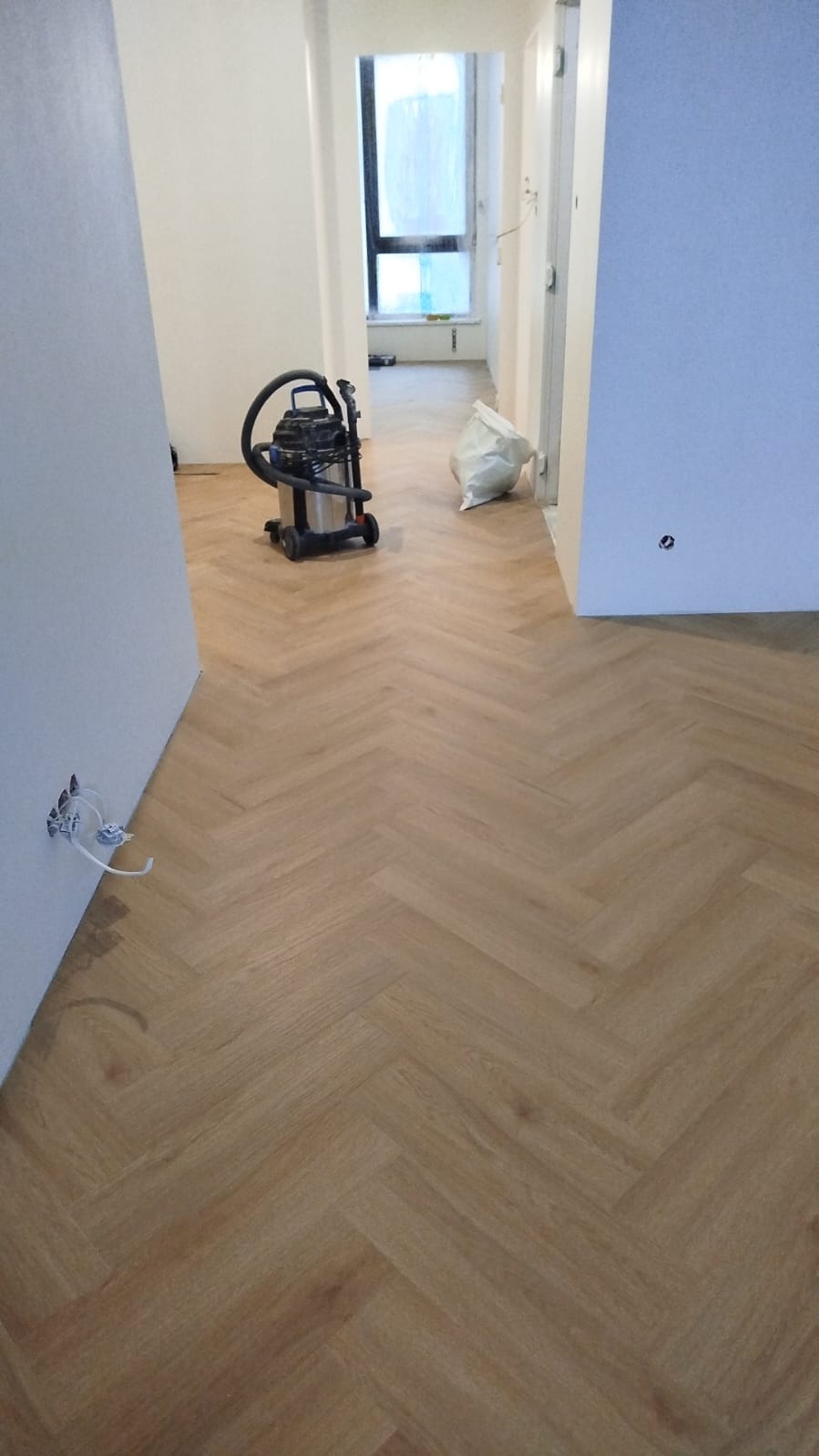 Ułożona podłoga w jodełkę, odkurzacz i worek z odpadami w tle. Widoczne gniazdko elektryczne bez osprzętu. Świeżo wykończone wnętrze w trakcie sprzątania.