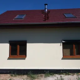 Dom jednorodzinny z nową, bordową dachówką ceramiczną, dwoma oknami dachowymi, kominem ze stali nierdzewnej i brązowymi oknami elewacyjnymi.