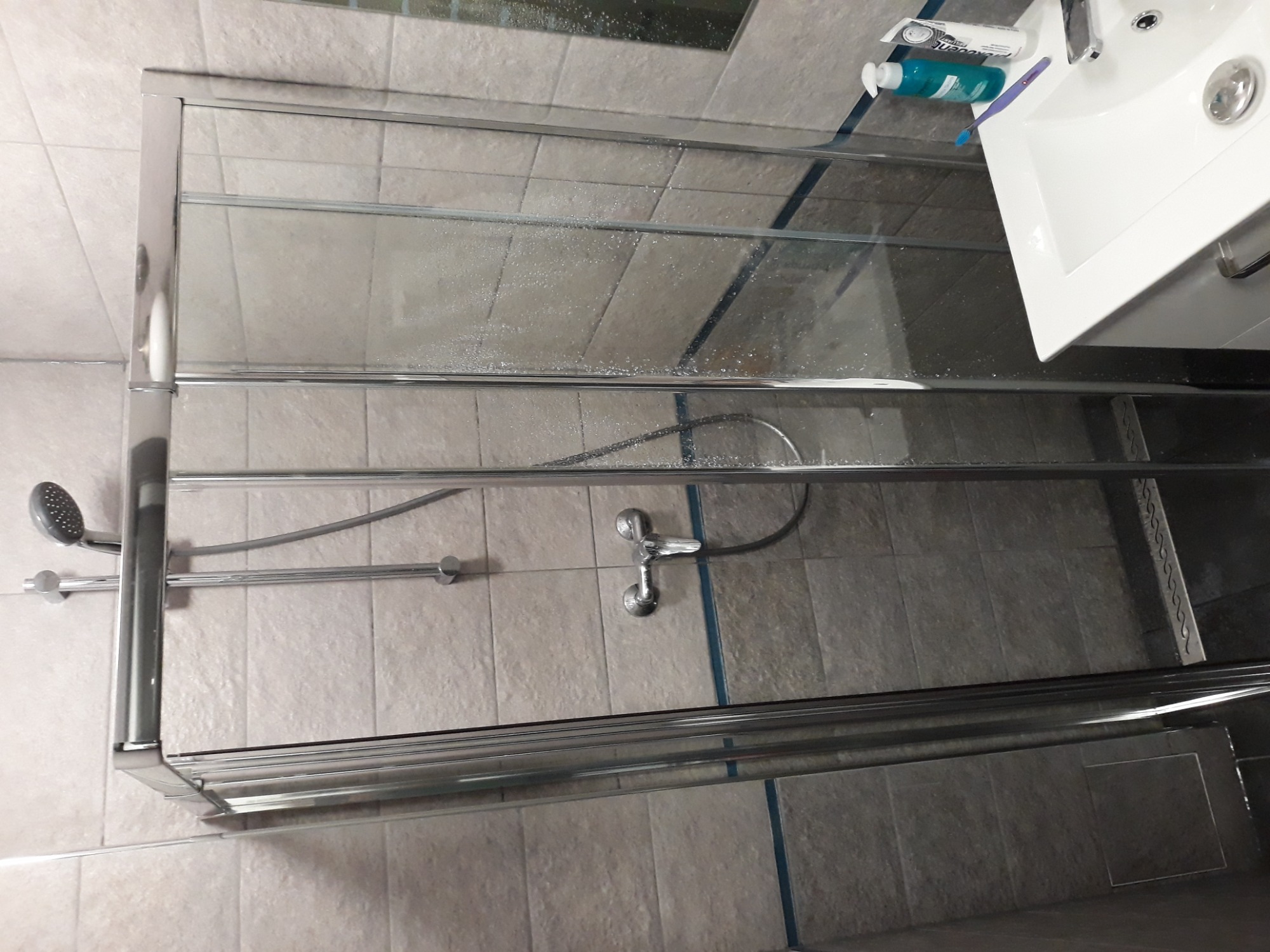 Szklana kabina prysznicowa z chromowanymi elementami, widoczna armatura, płytki ceramiczne na ścianach i podłodze, umywalka z kosmetykami w rogu, ujęcie z góry.