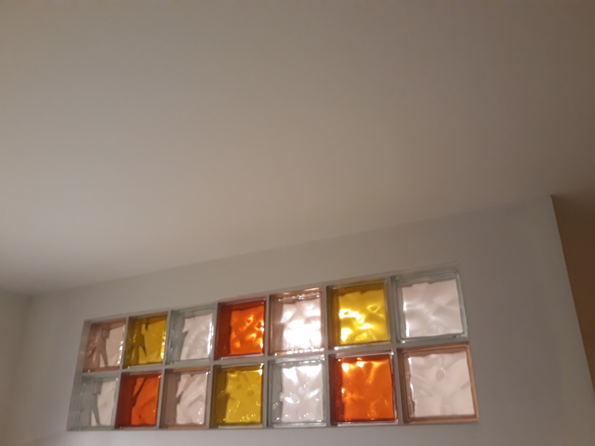 Ściana z wbudowanymi luksferami w odcieniach pomarańczy, żółci i bieli, widziana pod kątem od dołu, z fragmentem sufitu