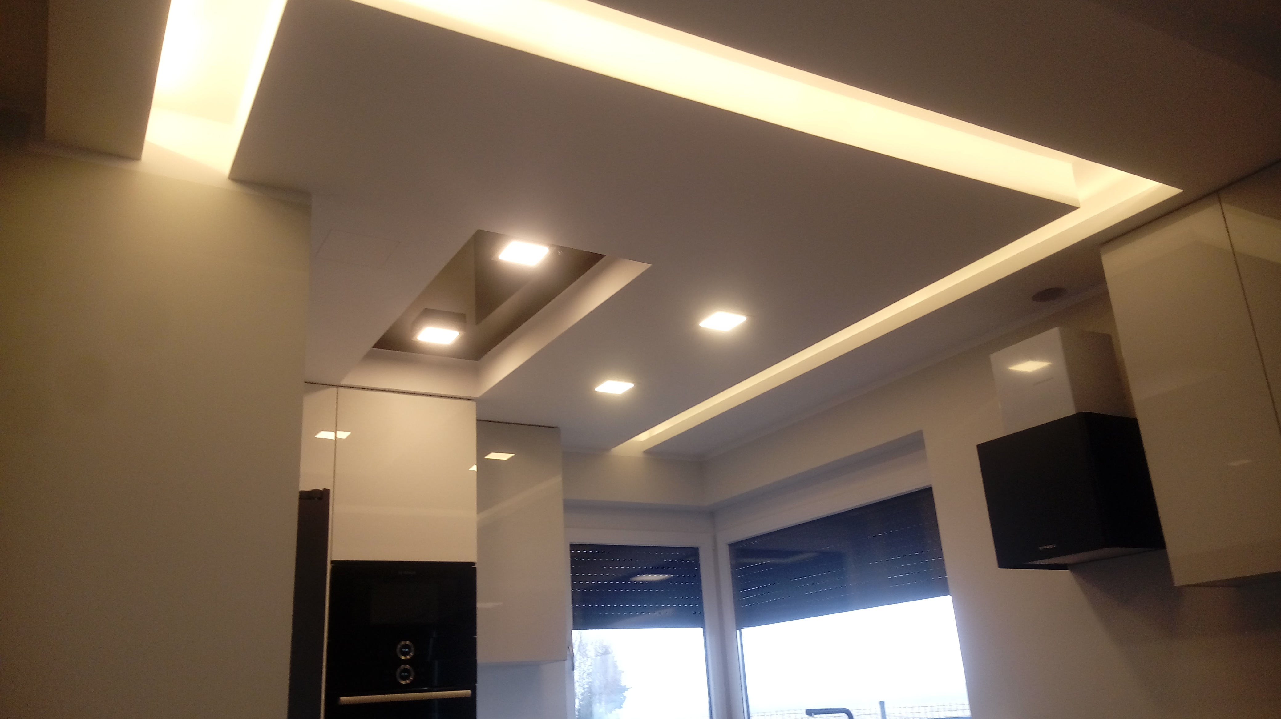 Nowoczesny sufit podwieszany z oświetleniem LED w kształcie prostokątów, widok z dołu, fragment zabudowy kuchennej z błyszczącymi frontami.