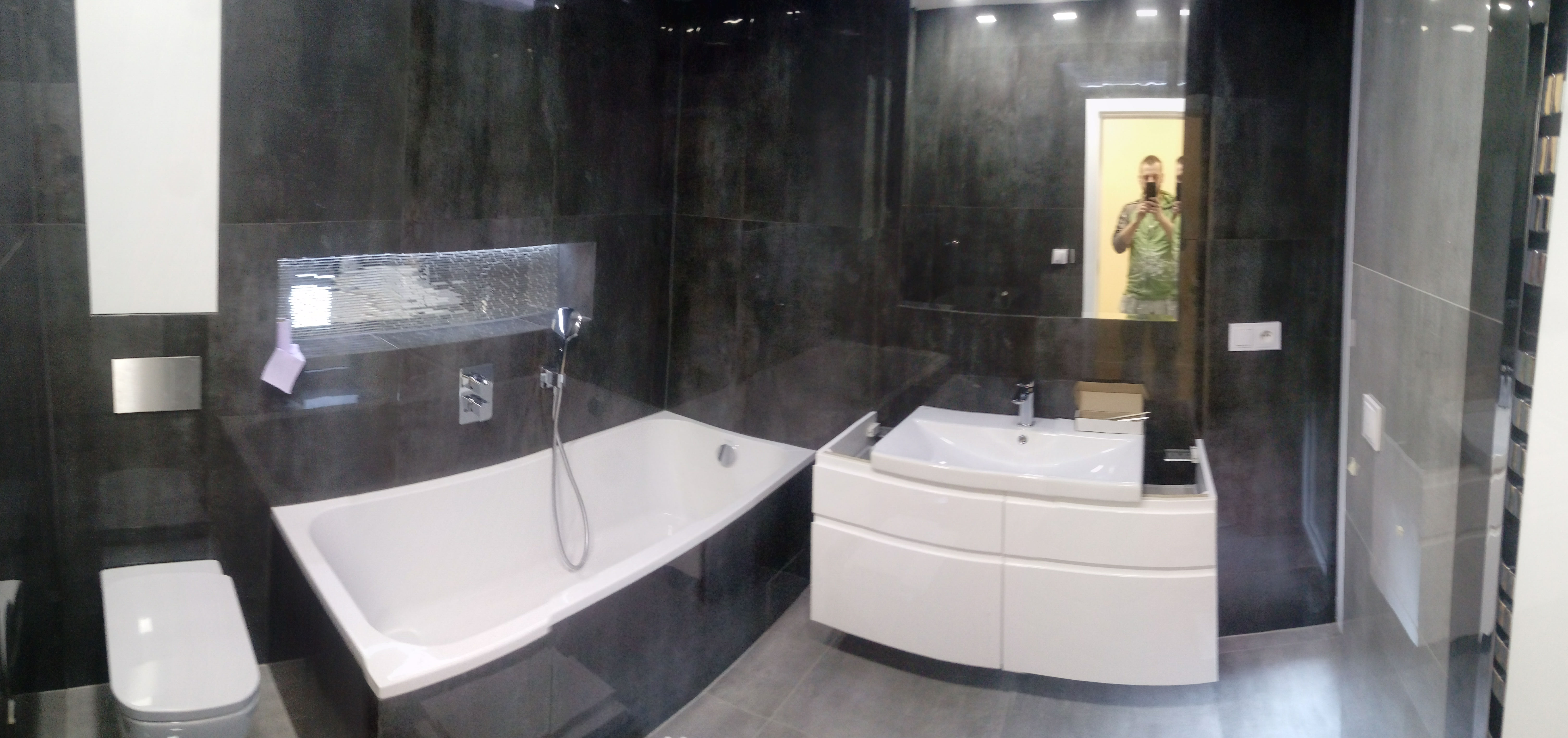 Nowoczesna łazienka z ciemnymi płytkami imitującymi kamień, białą wanną i umywalką z szafką, widoczne odbicie fotografa w lustrze.
