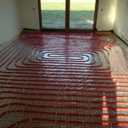 Czerwone rury układane spiralnie na folii izolacyjnej w jasnym pomieszczeniu z widokiem na zewnątrz przez duże okno balkonowe, przygotowanie do wylewki.