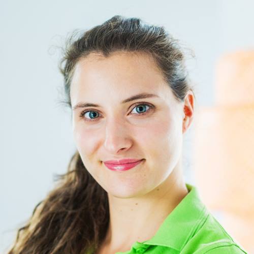 Portret uśmiechniętej kobiety o niebieskich oczach i brązowych włosach w zielonej koszulce, z rozproszonym, ciepłym światłem w tle.