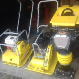 Żółte zagęszczarki gruntu i ubijak wibracyjny, modele Professional Line i Dro-Masz DRB80K, stoją obok siebie w pomieszczeniu.