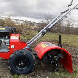 Czerwony glebogryzarek spalinowy na nierównym, lekko błotnistym terenie z widocznymi śladami pracy, tło stanowią drzewa i pochmurne niebo.