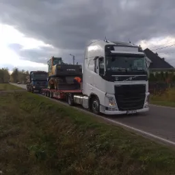 Dwa transportowe zestawy: biały ciągnik Volvo FH z naczepą przewozi żółtą koparkę Volvo, za nim jedzie ciężarówka MAN z przyczepą. Transport maszyn budowlanych.