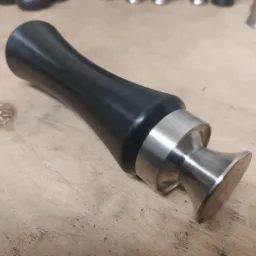 Precyzyjnie wykonany, dwuczęściowy element metalowy z czarną, gładką rękojeścią i srebrną, błyszczącą podstawą, leżący na drewnianym blacie z widocznymi śladami użytkowania.