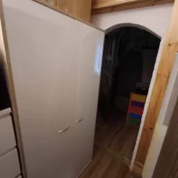 Biała szafa z połyskiem i drewnianym obramowaniem, umieszczona w pomieszczeniu z drewnianą boazerią i podłogą z paneli, widok na ciemne przejście z kolorową komodą.