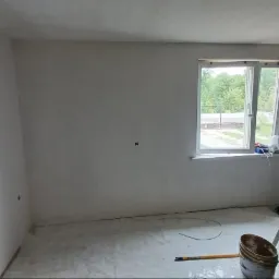 Wnętrze pokoju w trakcie prac wykończeniowych, ściany pokryte świeżą gładzią, widoczne narzędzia malarskie, drabina oraz fragment odsłoniętej ściany z cegły w sąsiednim pomieszczeniu.