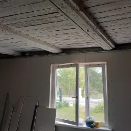 Pokój w trakcie remontu: ściany wyłożone płytami kartonowo-gipsowymi, sufit z drewnianymi belkami pokrytymi łuszczącą się białą farbą, okno z widokiem na zieleń, drabina i materiały budowlane...