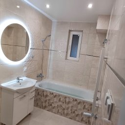 Roman Graczyk - Wykończona łazienka z beżowymi płytkami, podświetlanym okrągłym lustrem nad białą szafką umywalkową i wanną z dekoracyjną obudową.