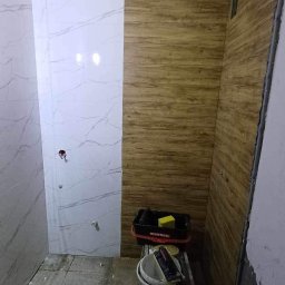 Roman Graczyk - Wnętrze łazienki w trakcie remontu: częściowo wyłożone płytkami imitującymi marmur, częściowo panelami ściennymi o wyglądzie drewna, widoczne narzędzia i wiadro na drabince.